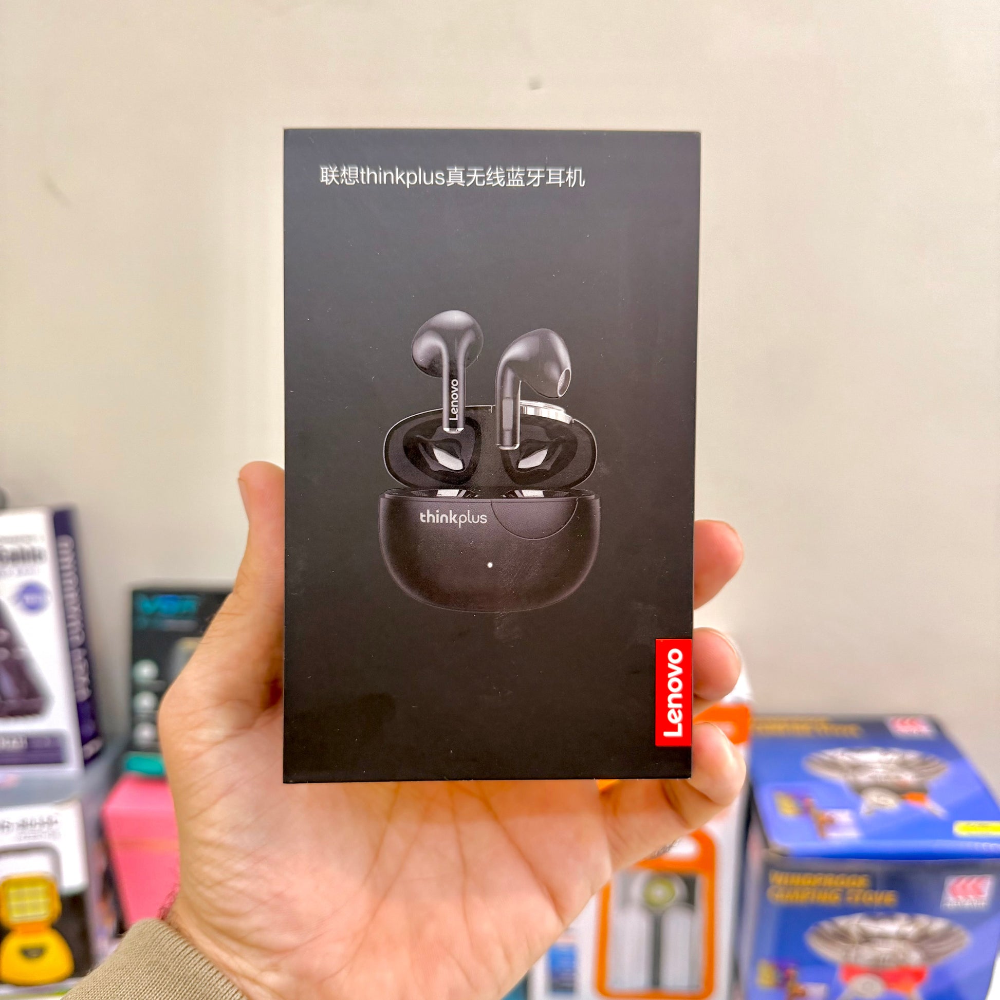 Hand holding a Lenovo Thinkplus wireless Bluetooth earphone box against a blurred indoor background